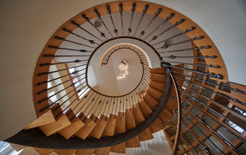 Ornate spiral stair case from below This architectural photograph shows an ornate spiral staircase taken from below in a building located in Devon, England, United Kingdom. The image captures the staircase’s elegant wooden steps and its decorative metal balustrade as it ascends in a circular formation towards the upper floors. Afternoon sunlight, typical of summer, illuminates the interior through windows, highlighting the warm tones of the wood and the surrounding walls. The view upward emphasizes the geometric design of the spiral staircase, while a contemporary chandelier hangs at the stairwell’s center, contributing to the refined ambience. This photograph exemplifies modern architecture and interior design, focusing on the details and craftsmanship of the staircase within an English setting.